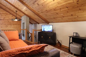 Ein gemütliches Zimmer mit Holzbalkendecke, einem Bett mit orangefarbener Bettwäsche, einem Fernseher auf einem schwarzen Standfuß und einer Treppe, die in ein Obergeschoss führt.