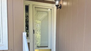 Unos escalones de madera conducen a una puerta con panel de vidrio, barandilla blanca y una lámpara fijada a la pared.