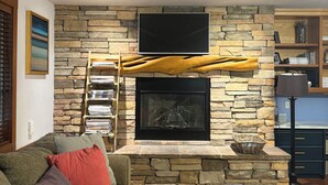 Una sala de estar con chimenea de piedra, un televisor de pantalla plana montado encima, una mesa de centro de madera y un sofá con cojines de colores.