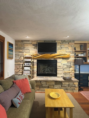 Una sala de estar con chimenea de piedra, un televisor de pantalla plana montado encima, una mesa de centro de madera y un sofá con cojines de colores.