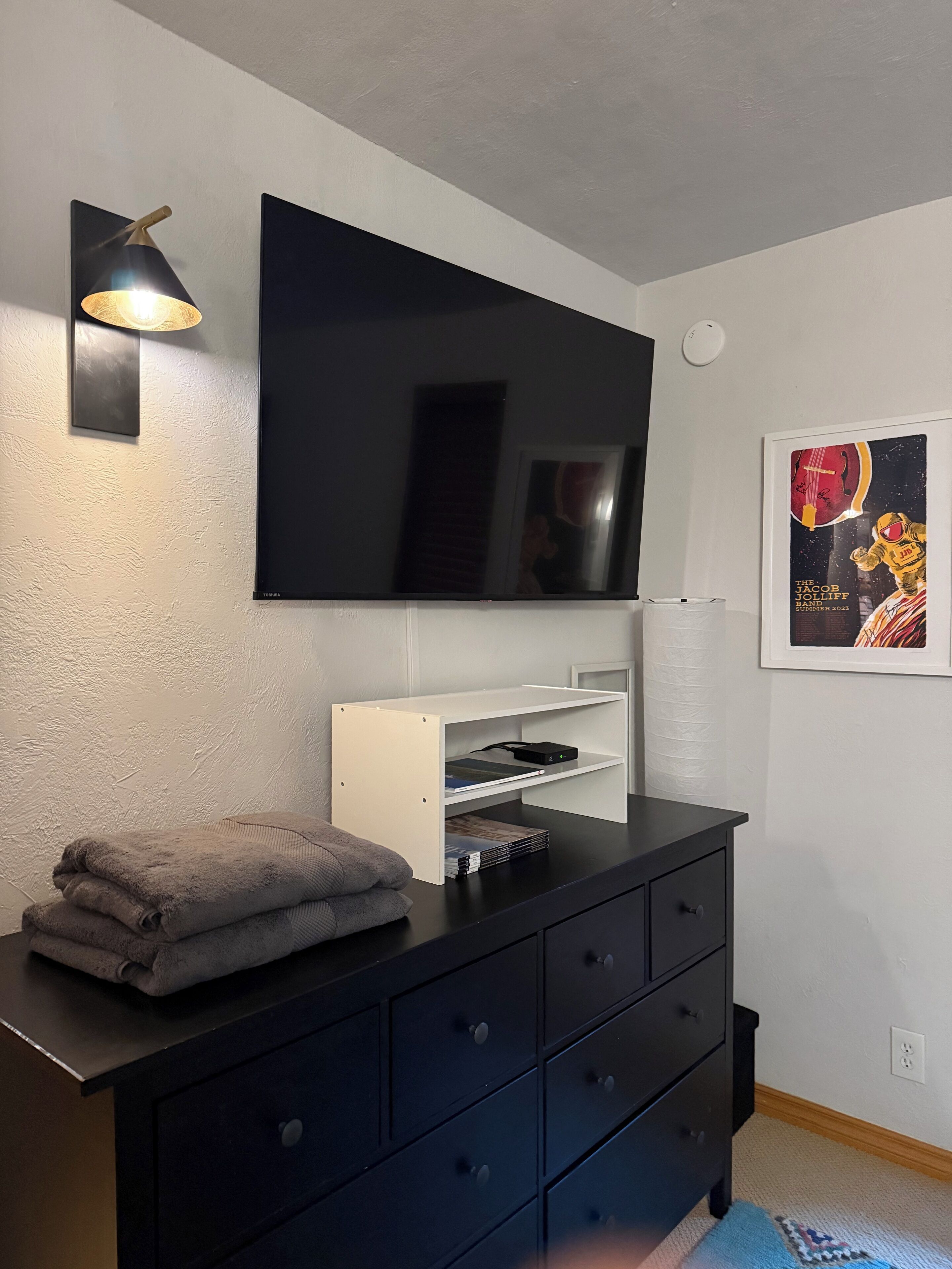 A modern room with a flat-screen TV mounted on the wall, a white shelf below, a black dresser with drawers, and a framed poster on the wall.