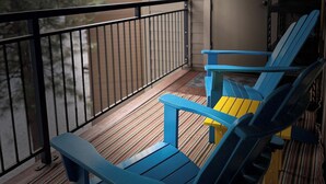 Un balcón con dos sillas Adirondack azules y una silla amarilla, baranda negra, y vista a una casa y árboles.