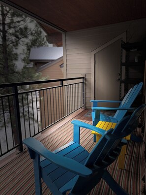 Un balcón con dos sillas Adirondack azules y una silla amarilla, baranda negra, y vista a una casa y árboles.