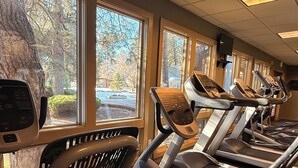 Un gimnasio con cintas de correr y bicicletas estáticas, ventanas amplias con vista a árboles y un estacionamiento.