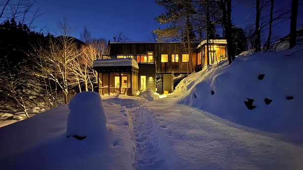 A snow-covered path leading to a modern house with multiple levels and large windows, surrounded by trees.
