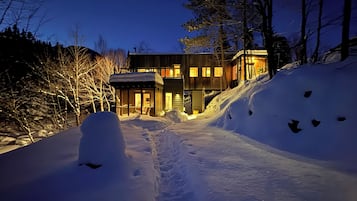 Un sentier enneigé mène à une maison moderne à plusieurs niveaux, dotée de grandes fenêtres et entourée d’arbres.