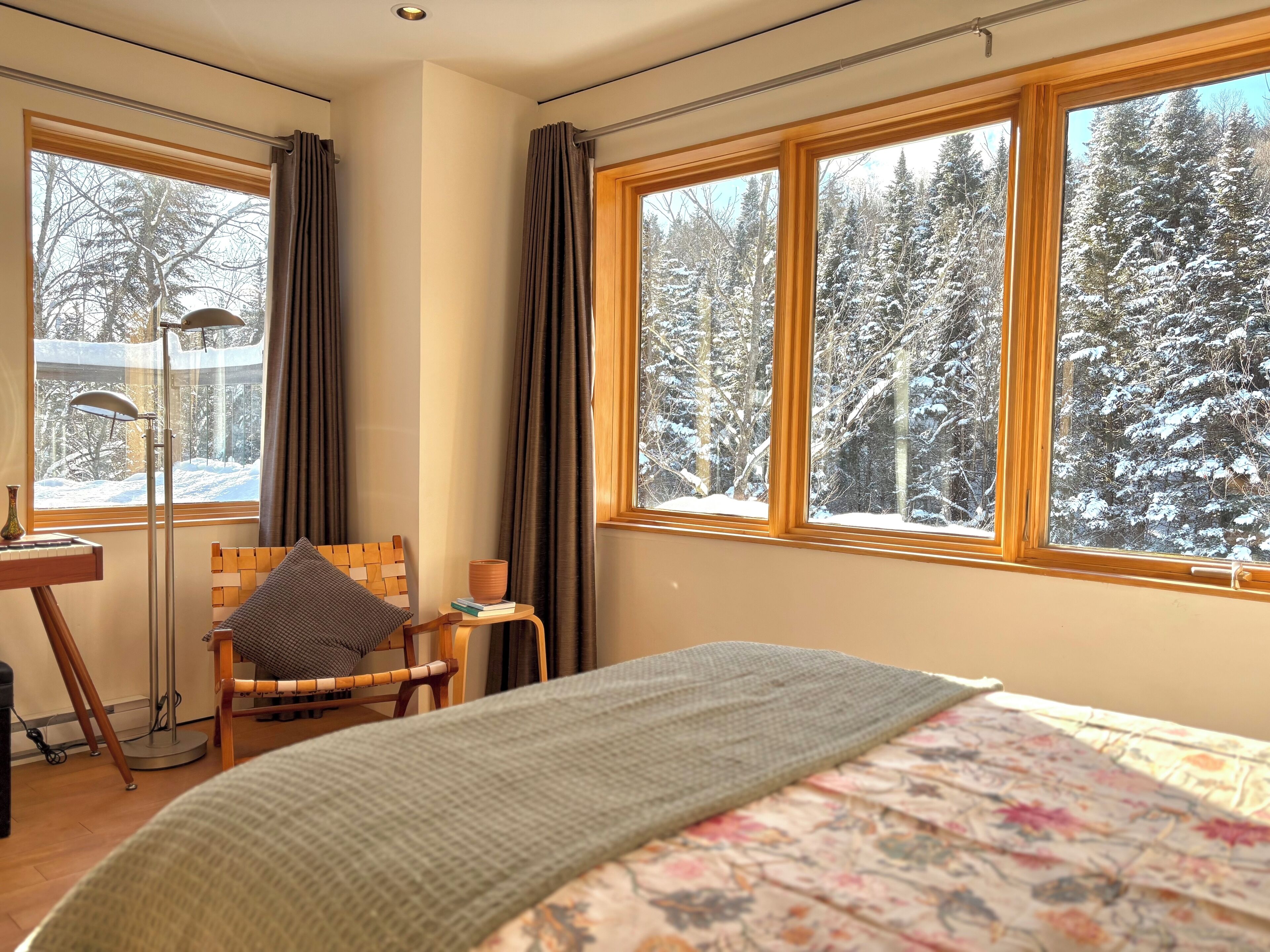 Una habitación acogedora con una cama, una silla, una mesita con una taza y grandes ventanales que dan a un bosque nevado.