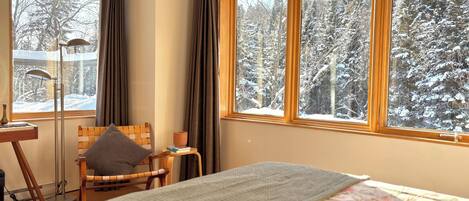 Una habitación acogedora con una cama, una silla, una mesita con una taza y grandes ventanales que dan a un bosque nevado.