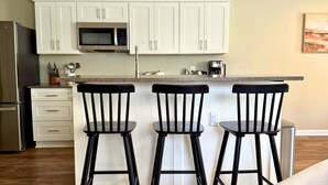 A kitchen with white cabinets, a microwave, and a coffee maker. There are three black bar stools and a stainless steel refrigerator.