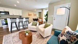 A modern living room with a sofa, a coffee table, and a patterned rug. There are two chairs and a dining area with a table and chairs in the background.