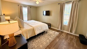 A bedroom with a bed, a nightstand with a lamp, a ceiling fan, and a TV on the wall.