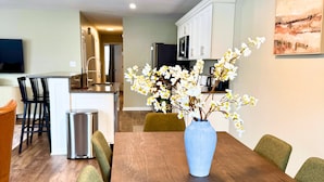 A dining area with a wooden table, chairs, and a vase with flowers. There is a kitchen in the background with cabinets and appliances.