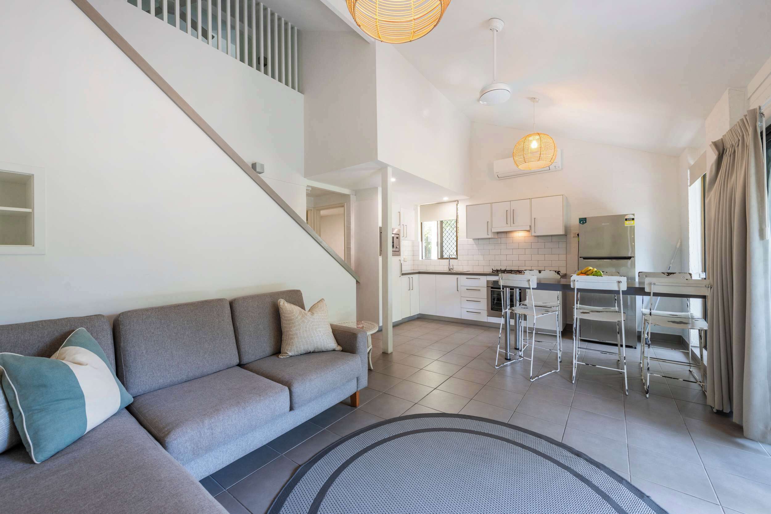 A modern apartment with a grey sofa, a staircase, a kitchen with white cabinets, and a dining area with metal chairs.