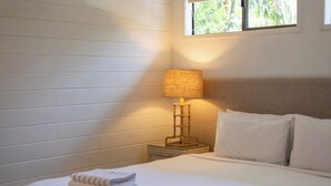 A neatly made bed with white linens and two pillows, a bedside lamp on a small table, and a window with a view of greenery.