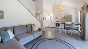 A modern apartment with a grey sofa, a staircase, a kitchen with white cabinets, and a dining area with metal chairs.