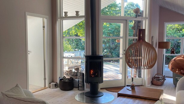 A modern living room with a central wood-burning stove, large windows, and a cozy seating area. - Holiday oasis with terrace and sloping garden (Tikøb)