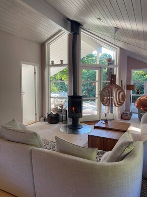 A modern living room with a central wood-burning stove, large windows, and a cozy seating area. - Holiday oasis with terrace and sloping garden (Tikøb)