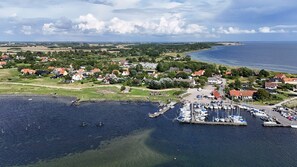 Vista aérea de un pueblo costero con un puerto deportivo, casas y aerogeneradores en la distancia.