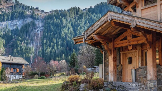 A wooden cabin with a stone foundation, surrounded by a grassy area and a forested mountain backdrop.