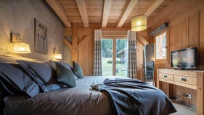 A bedroom with a wooden ceiling, a bed with pillows, a bedside lamp, a TV on a wooden dresser, and a view of the outdoors through a glass door.