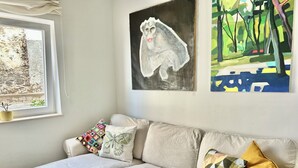 Een woonkamer met een beige bank, een raam met uitzicht op een stenen muur, en drie kunstwerken aan de wand.