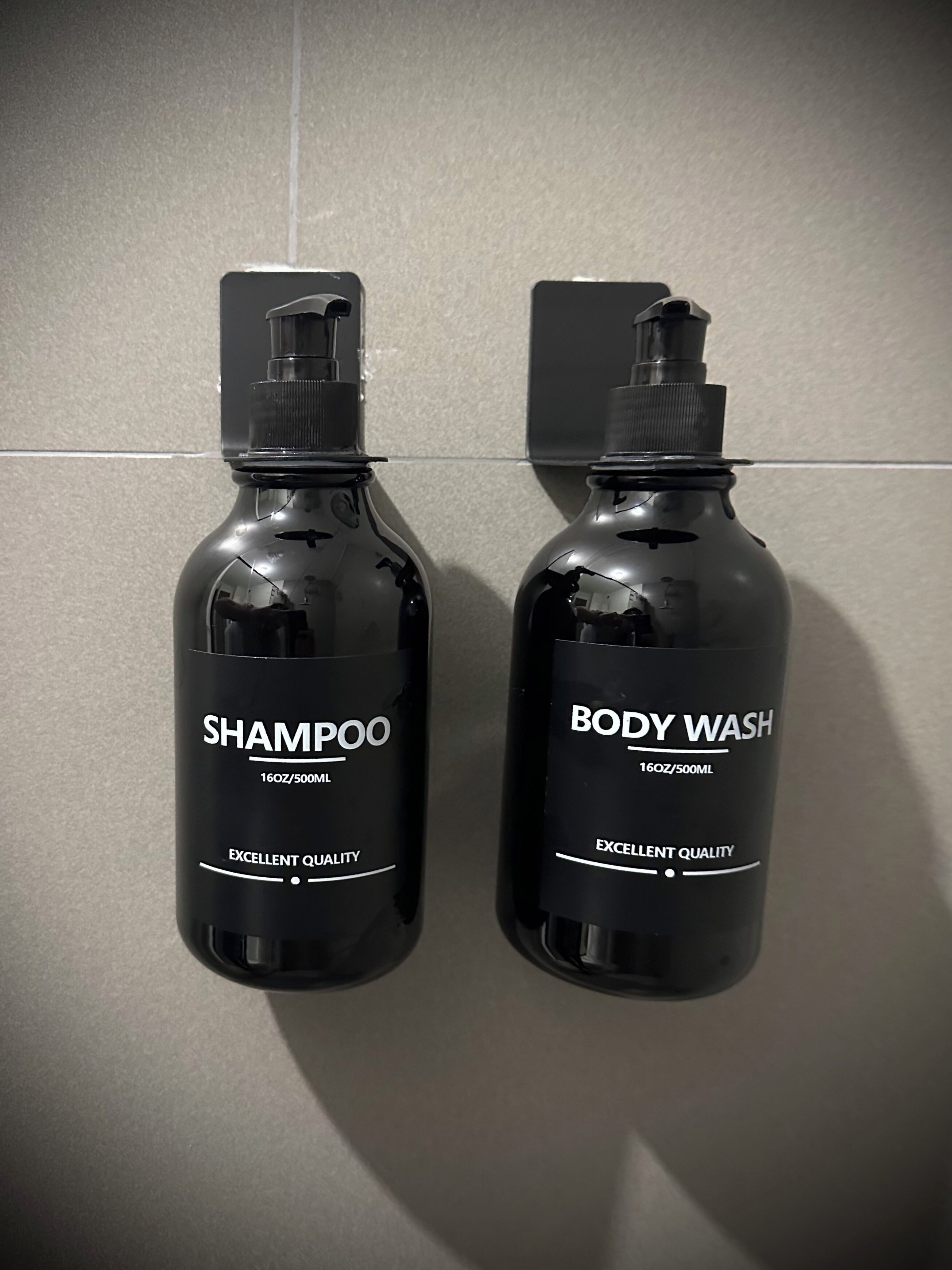 Kaksi mustaa shampoopulloa ja pullo suihkugeeliä laattaseinällä.