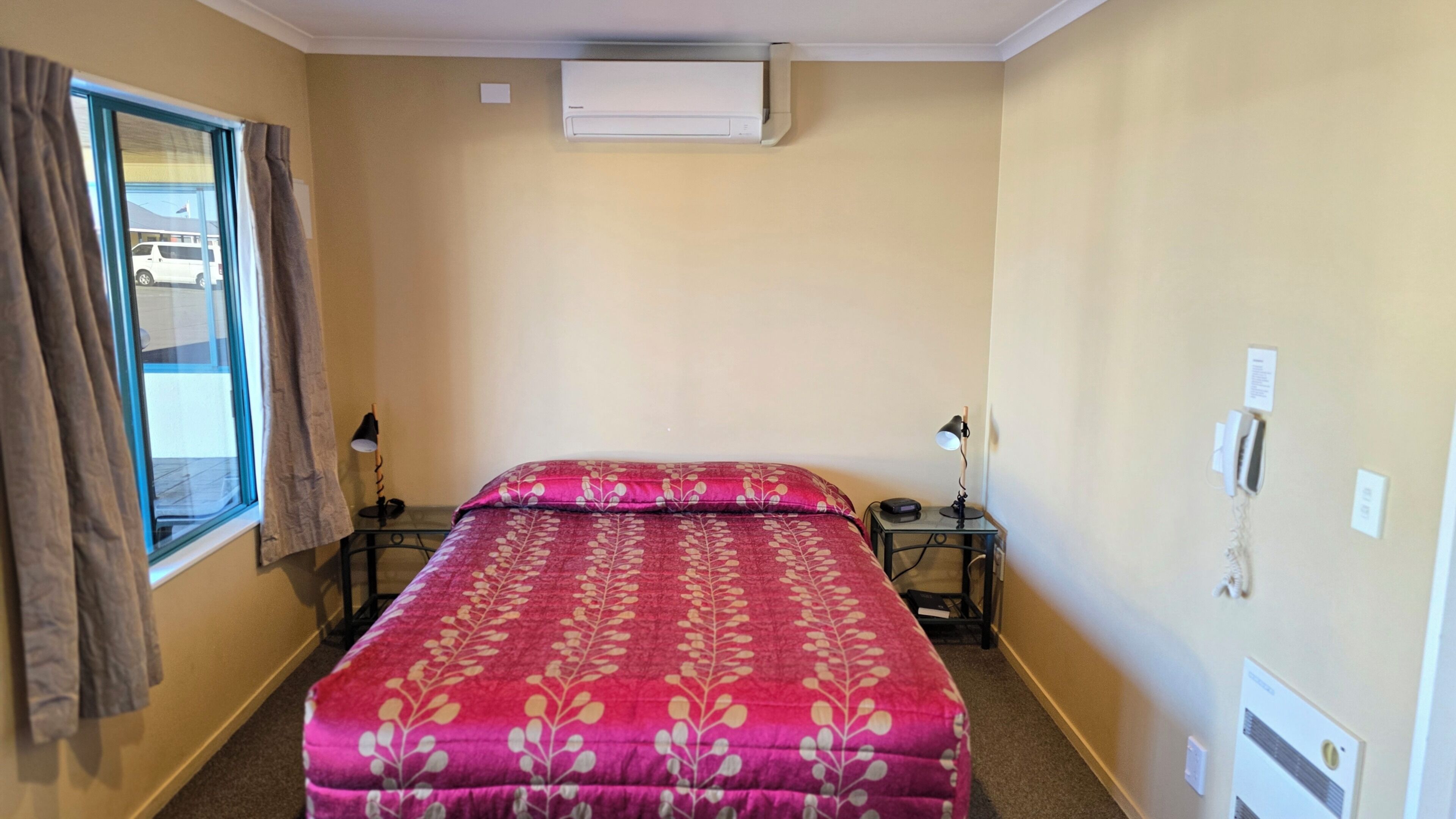 Habitación de hotel con cama, dos mesitas de noche, ventana con cortinas, equipo de aire acondicionado y teléfono instalado en la pared.