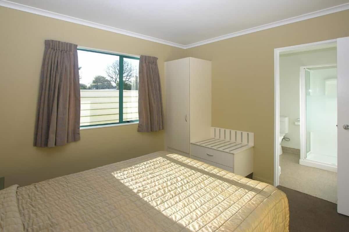 Un dormitorio con cama, armario, ventana con cortinas y puerta que da a un baño.