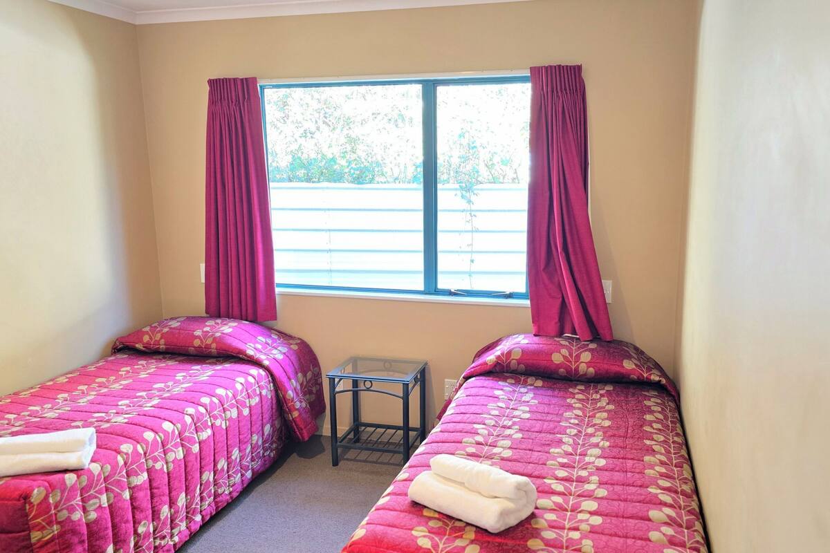 Una habitación con dos camas, cada una con una colcha de flores rosadas y una toalla blanca. Hay una mesita entre las camas y una ventana con cortinas.