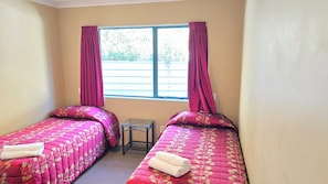 Una habitación con dos camas, cada una con una colcha de flores rosadas y una toalla blanca. Hay una mesita entre las camas y una ventana con cortinas.