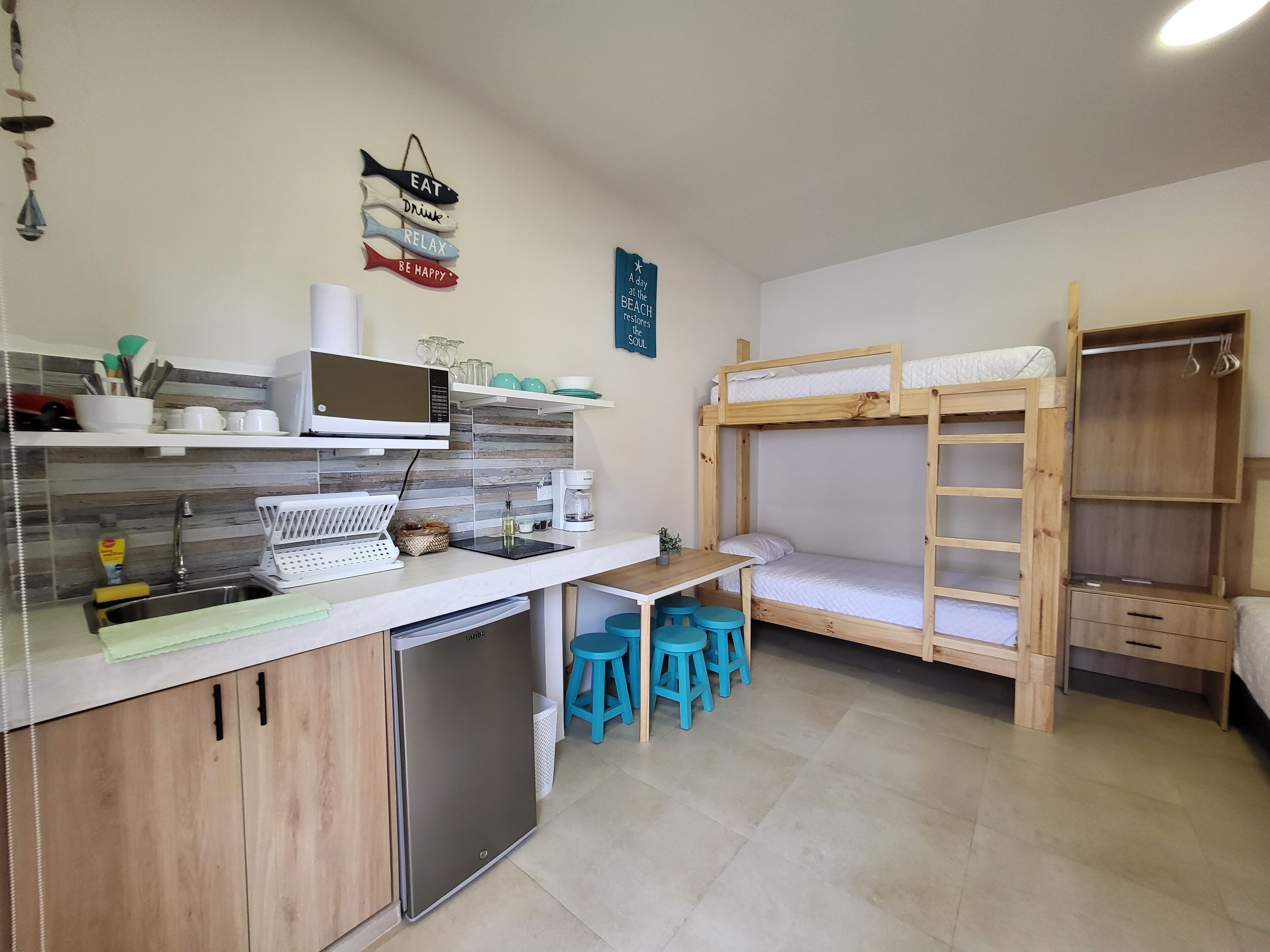 A compact kitchen with a microwave, sink, and a small refrigerator. There are stools and a bunk bed with a ladder.