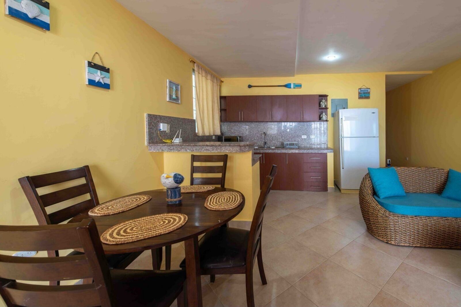 Una cocina compacta con armarios de madera, una mesa de comedor con sillas y un sofá de mimbre con cojines azules.