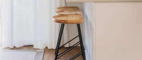 Una cocina con encimera blanca, armarios de madera, un taburete de metal negro y una cortina blanca.