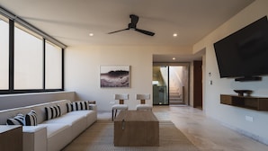 A modern living room with a sofa, a coffee table, a ceiling fan, a flat-screen TV, and a large window. - Penthouse Arenamar in Punta Mita (Punta de Mita)
