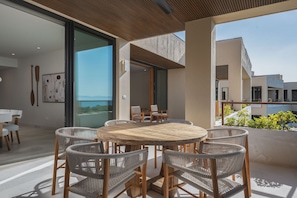 An outdoor dining area with a round wooden table and wicker chairs, large windows, and a view of buildings. - Penthouse Arenamar in Punta Mita (Punta de Mita)