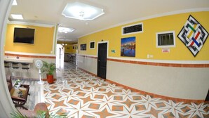 Un couloir d’hôtel avec un revêtement de sol à motifs, un téléviseur fixé au mur et une plante en pot.