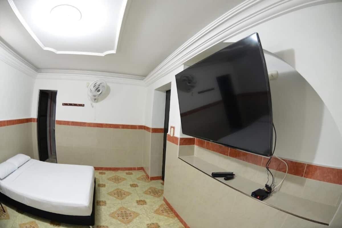 Une petite chambre d’hôtel comprenant un lit, un téléviseur à écran plat fixé au mur, un ventilateur de plafond et un sol carrelé.