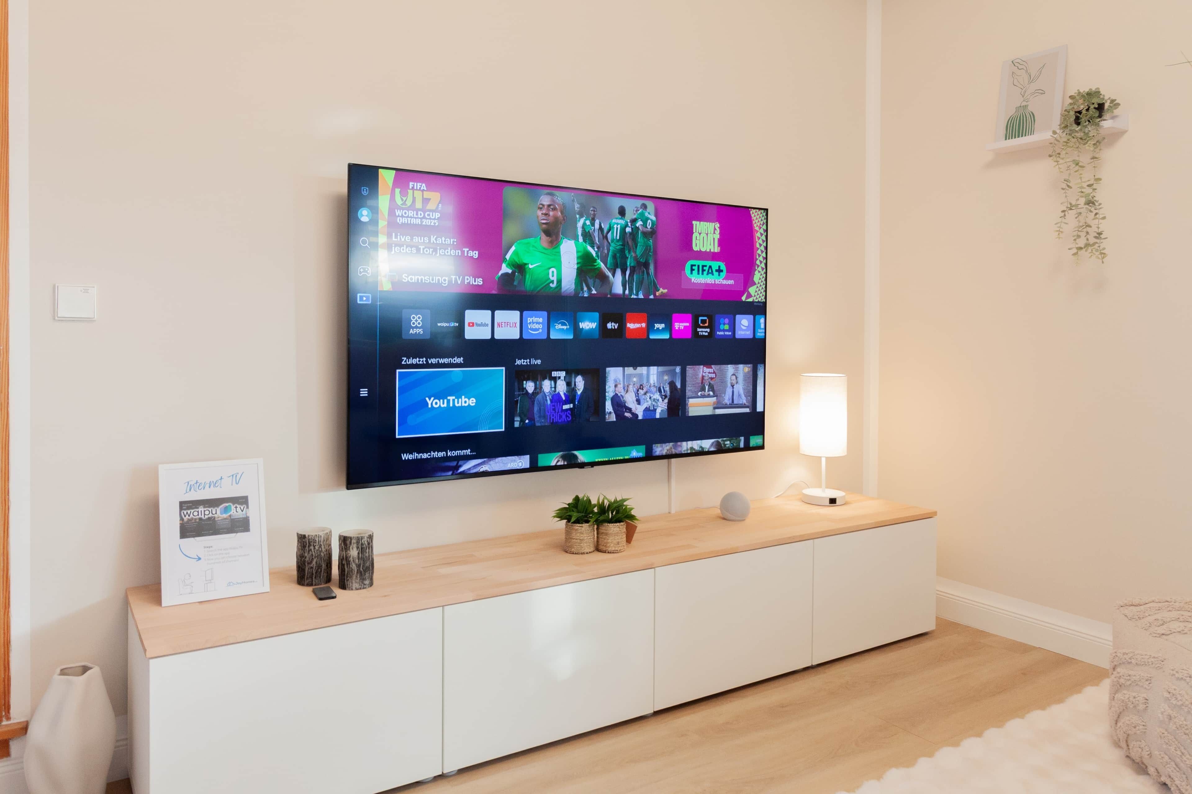 A modern living room with a flat-screen TV mounted on the wall, a white entertainment unit below, and a small table with a lamp and decorative items.