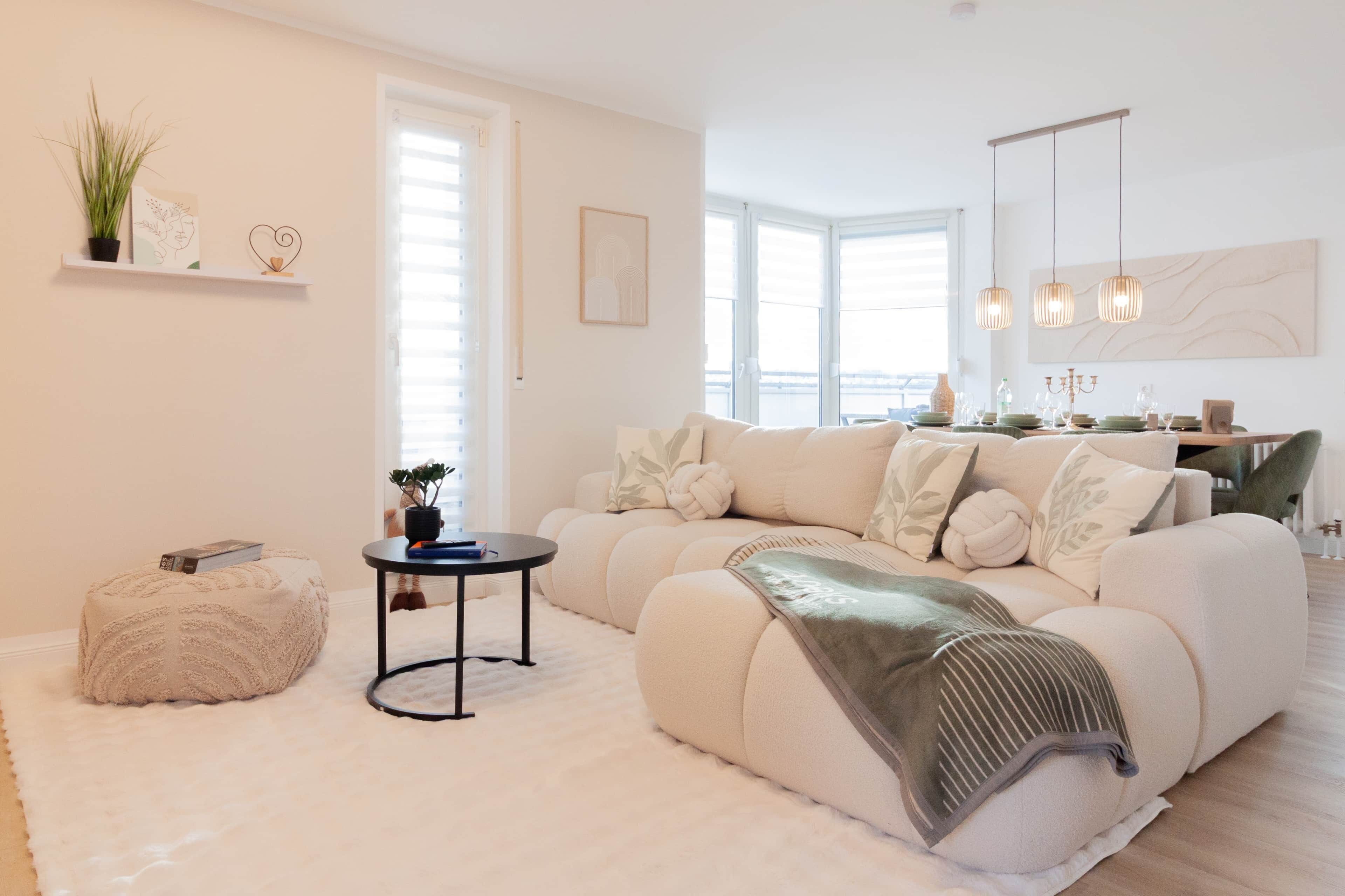 A modern living room with a sectional sofa, a round coffee table, and a small ottoman. There is a dining area with a table set for four, and a large window providing natural light.