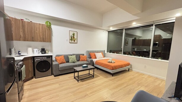 A compact living space with a sofa, a bed, a coffee table, a washing machine, and a large window offering a view of the kitchen. - 12th Floor Studio at Downtown.  Parking Included (Edmonton)