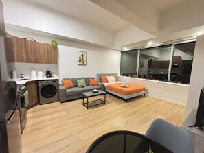 A compact living space with a sofa, a bed, a coffee table, a washing machine, and a large window offering a view of the kitchen. - 12th Floor Studio at Downtown.  Parking Included (Edmonton)
