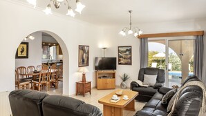 Ein Wohnzimmer mit Sofa, Fernseher, Esstisch und Stühlen. Es gibt einen Fernsehschrank, eine Lampe und ein Bild an der Wand.