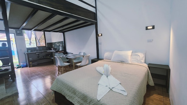 Un dormitorio con cama, escritorio y silla. Hay una ventana con cortinas, una puerta y un interruptor de luz en la pared.