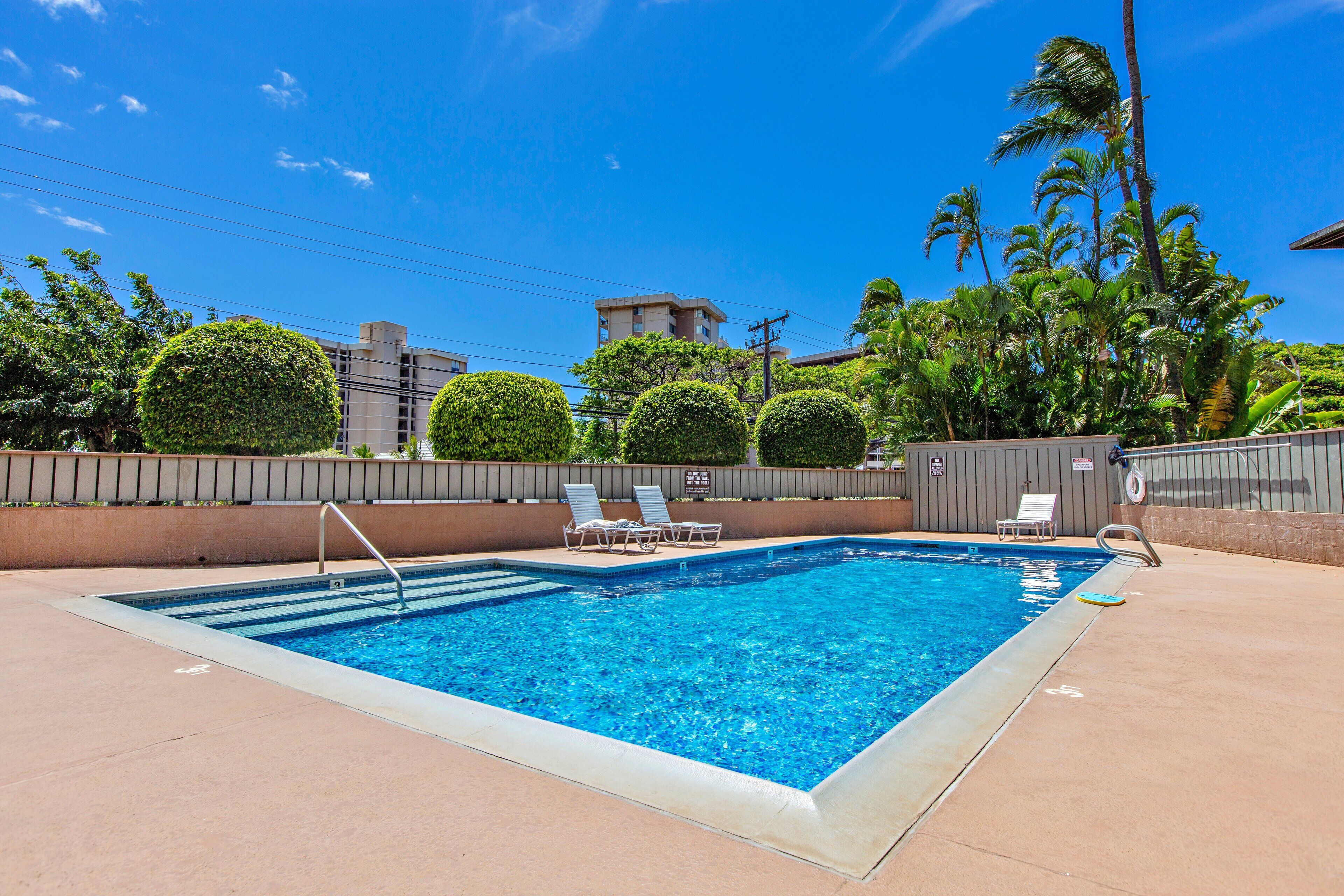 A rectangular swimming pool with a clear blue water, surrounded by a low brick wall. There are lounge chairs along the poolside, and a lifeguard stand is visible.
