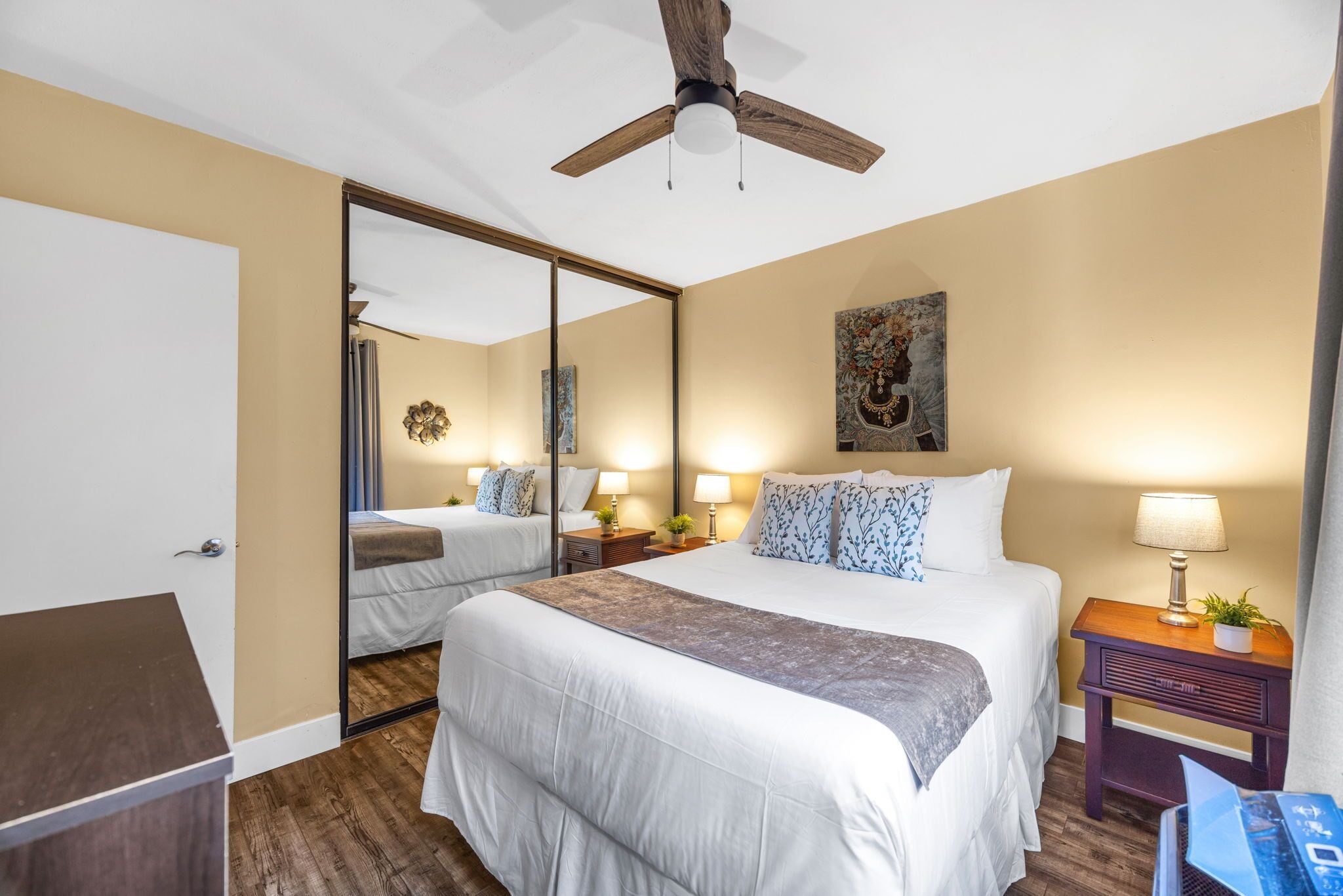 Una habitación de hotel con una cama grande, un ventilador de techo, un espejo y una mesita de noche con lámpara.