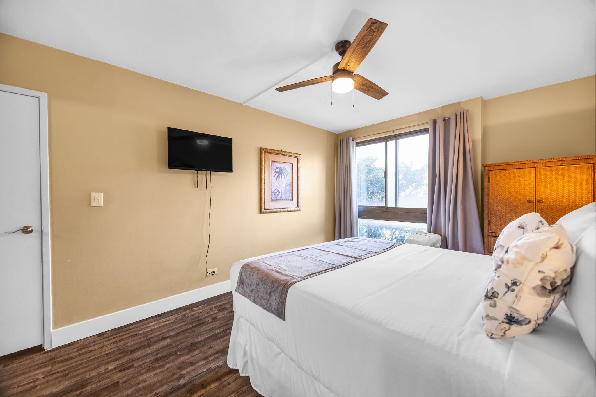Quarto de hotel com cama, ventoinha no teto, televisão, janela com cortinas e guarda-roupa de madeira.
