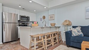 A modern kitchen with stainless steel appliances, a wooden island with stools, and a blue sofa in a living area. - Oceanfront Getaway with Stunning Views , Pool, & Easy Beach Access (North Myrtle Beach)