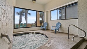 A spa area with a hot tub, two blue Adirondack chairs, and large windows offering a view of palm trees and a building. - Oceanfront Getaway with Stunning Views , Pool, & Easy Beach Access (North Myrtle Beach)