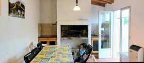 Uma área de refeições com uma mesa coberta por uma toalha colorida, cadeiras pretas, um lustre pendente e vista para o exterior através de uma porta de vidro.