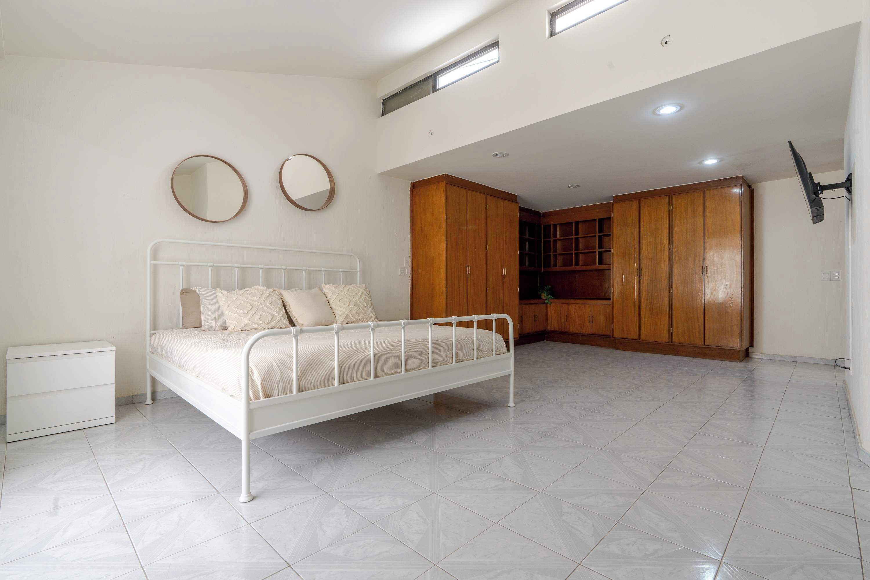 A bedroom with a white metal bed, a white nightstand, wooden wardrobe, and a small window.